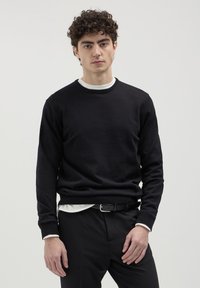 Giovane con capelli ricci che indossa un maglione nero sopra una camicia bianca, pantaloni neri e una cintura nera, in piedi su uno sfondo semplice.