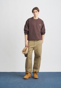 Hanorac maro, pantaloni largi khaki, cizme bej și o șapcă bej. Materialele par moi, dar durabile; designul este relaxat și practic.