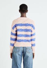 Personne aux cheveux courts et bouclés portant un pull duveteux rayé rose et bleu ainsi qu'un jean en denim bleu, debout de dos sur un fond blanc.