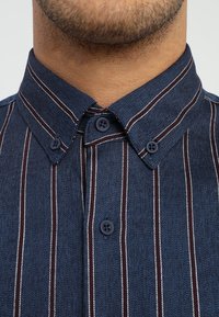 Närbild av en mans hals och haka i en blå skjorta med knappar och vita och mörkr öda vertikala ränder.