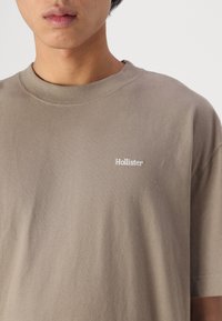 Grijze katoenen t-shirt met een ronde hals en korte mouwen. Bevat een klein wit "Hollister" logo op de linkerkant van de borst. Zachte textuur.