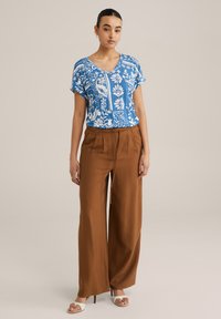 Zila ziedu apdrukas īsām piedurknēm augšdaļa ar V veida kakla izgriezumu, savienota ar brīvi krītošiem, augstā vidukļa brūniem biksēm, kas izgatavotas no viegla auduma.