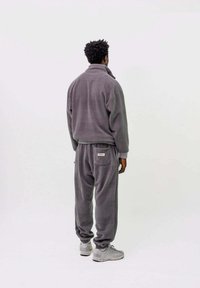 Grauer Fleece-Trainingsanzug mit hohem Kragen und elastischen Bündchen, ausgestattet mit einer Rückentasche. Getragen mit grauen Turnschuhen.