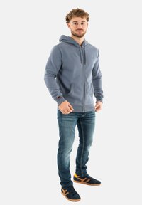 Grijze rits-hoodie met een capuchon, voorzakken en geribbelde manchetten; gecombineerd met blauwe jeans en zwarte sneakers met oranje accenten.