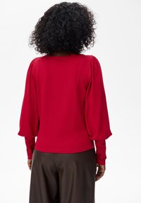 Osoba z kręconymi włosami ubrana w czerwoną bluzkę z długimi rękawami i bufkami oraz brązowe spodnie, pokazana od tyłu na białym tle.