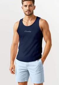 Navyblaues Tanktop mit rundem Ausschnitt und weißem Schriftzug "Paradise" auf der Vorderseite. Kombiniert mit hellblauen Shorts.