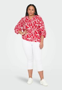 Röd och vit mönstrad blus med en V-hals, långa puffärmar, tillsammans med vita cropped byxor och vita sneakers mot en ljus bakgrund.