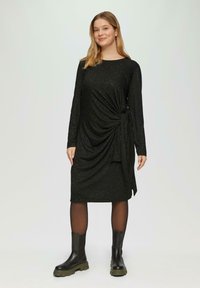 Schwarzes Kleid aus strukturiertem Strick, mit langen Ärmeln und einem verdrehten Frontdesign mit einer Schleifendetail an der Seite. Getragen mit schwarzen Ankle Boots.