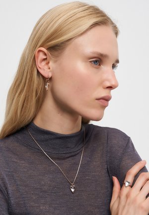 Donna bionda in top scuro trasparente che indossa una collana con pendente a forma di cuore, anello e orecchini coordinati, che guarda pensierosa di lato.