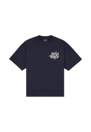 Femme portant un t-shirt oversize bleu marine avec un texte blanc, une mini jupe en cuir noire, de grandes bottes noires, des lunettes de soleil et tenant un sac à main noir.