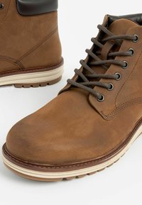 Bruine leren enkelboots met een donkerbruine kraag, ronde neus en grijze veters. Voorzien van een witte rubberen zool en subtiele stikseldetails.
