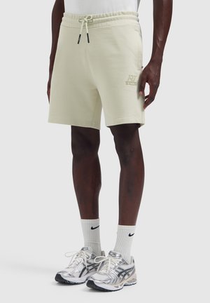 Mann trägt beige Shorts mit Kordelzug und Logo, weiße Nike-Socken und silberne Sportschuhe, steht vor schlichtem Hintergrund.