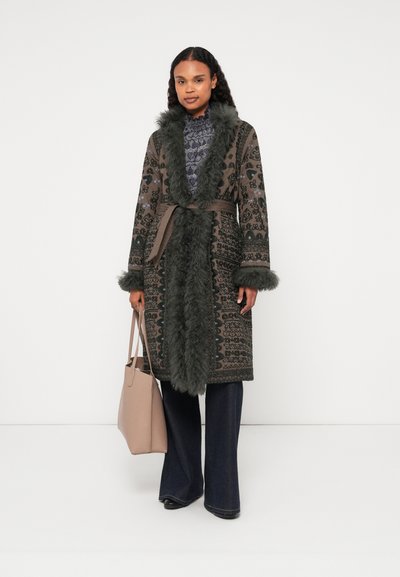 Μακρύ παλτό με σχέδιο σε σκούρο πράσινο χρώμα με τεχνική γούνα, ζώνη στη μέση. Το μοντέλο κρατάει μία ανοιχτή μπεζ τσάντα, φορώντας σκούρες φαρδιές τζιν.