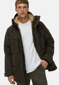 Dunkelbraune isolierte Jacke mit Kapuze, ausgestattet mit einem gelben Fleece-Futter, mehreren vorderen Taschen und einem Reißverschluss. Begleitend dazu ein weißes Shirt.
