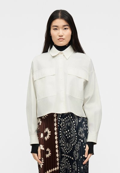 Jeune femme aux longs cheveux bruns portant une chemise crème boutonnée surdimensionnée par-dessus des manches noires et une jupe plissée à motifs floraux marron et marine.