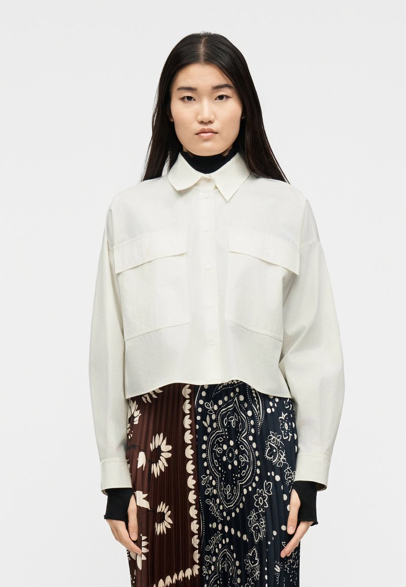 Νεαρή γυναίκα με μακριά σκουρόχρωμα μαλλιά φορά ει ένα φαρδύ κρεμ πουκάμισο με κουμπιά πάνω από μαύρα μανίκια και μια πλισέ φούστα με καφέ και ναυτικό μπλε λουλουδάτα σχέδια.