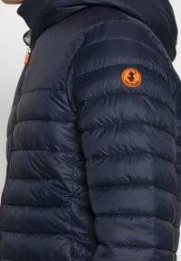 Mörkblå vadderad jacka med dragkedja, med horisontell sömnad, en orange cirkulär logotyp på vänster ärm och sidofickor.