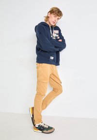 Hoodie marine avec détails en patch, pantalon cargo beige, et baskets multicolores. Accent sur le confort avec une coupe décontractée.