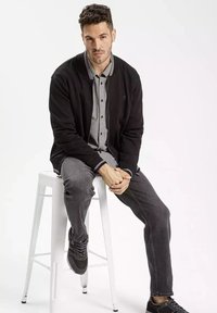 Czarny kardigan nałożony na koszulę w kratę, w połączeniu z ciemnymi dżinsami i czarnymi sneakersami. Model siedzi na białym stołku, z rękami splecionymi.