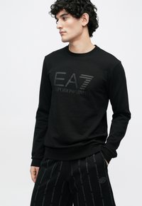 Fiatalember göndör hajjal, fekete EA7 Emporio Armani pulóvert és hozzáillő márkázott nadrágot viselve, oldalra néz, kezét zsebre téve.