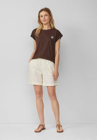 Woman stands against gray wall wearing brown short-sleeve top with embroidered turtle, beige shorts, and brown sandals, hands in pockets.