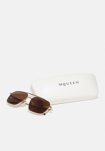 Gafas de sol marrones con un diseño de montura geométrica, detalles en oro y un estuche duro blanco con el nombre de la marca "MQUEEN" impreso en él.