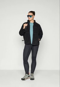 Must tume fleece-jakk kahe eesmine taskuga, helesinine särk, mustad leggingud küljetaskutega ja hallid matkasaapad. Sinise varjundiga päikeseprillid.