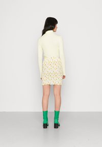 Suéter justo amarelo claro com gola alta combinado com uma saia mini branca com padrão floral. A modelo usa botas de salto verdes. Tecido suave e detalhes texturizados.