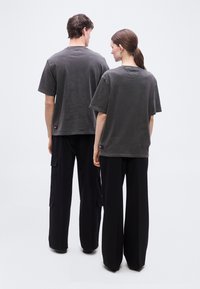 Δύο άτομα που στέκονται με την πλάτη προς τα εμπρός, φοράνε γκρι φαρδιές μπλούζες και μαύρο παντελόνι με φαρδιά μπατζάκια, μπροστά σε απλό φόντο.