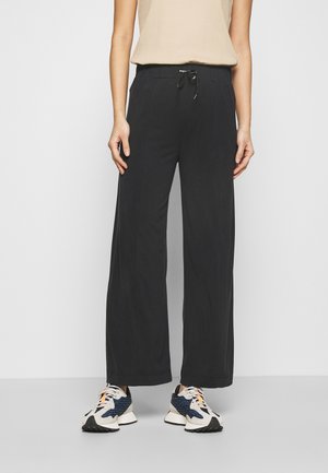 Woman wearing black wide-leg pants with a beige sleeveless top and white, blue, and orange sneakers, standing against a plain background.