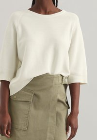 Pull crème à col large et manches trois-quarts, associé à une jupe khaki avec une poche latérale et une ceinture.