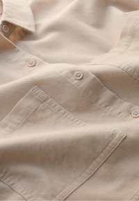 Beige button-up shirt with a textured fabric, featuring two buttons and a chest pocket detail. Soft, wrinkled appearance.