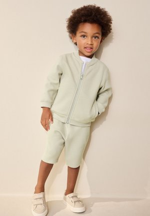 Kind met krullend haar dat een lichtgroen jack met rits draagt, bijpassende korte broek en beige klittenbandsneakers, staande tegen een beige muur.