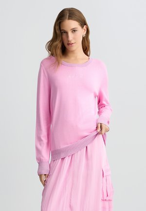 Maglione rosa in morbido tessuto, con maniche lunghe, collo rotondo e orlo a coste con taglio asimmetrico. Dettaglio del logo visibile.