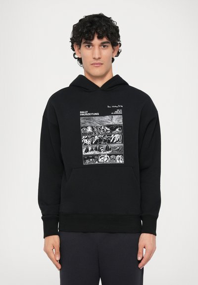 Man wearing black hoodie with white graphic panels depicting outdoor scenes and text "BALLY HAUSZEITUNG" on front, standing against white background.