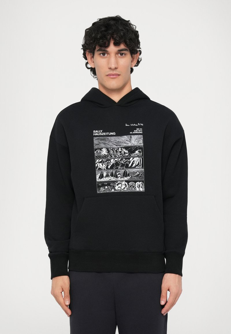 Uomo che indossa un hoodie nero con pannelli grafici bianchi che raffigurano scene all'aperto e il testo "BALLY HAUSZEITUNG" sulla parte anteriore, in piedi contro uno sfondo bianco.