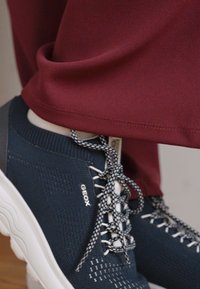 Marinblå sneaker med texturerad stickad ovandel, vit sula och mönstrade snörningar. Bärs med vinröda byxor, vristerna synliga. Geox-logotyp synlig.