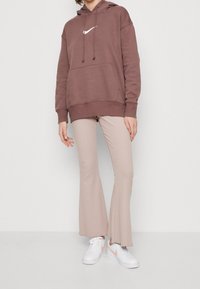 Person med en brun Nike-hoodie, beiga ribbade flarebyxor och vita sneakers med persiko-Nike-swoosh, stående framför en enfärgad bakgrund.