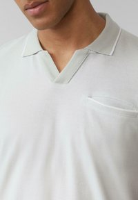 Lichtgrijze poloshirt met een v-hals en contrasterende kraag. Voorzien van een klein borstzakje en een gestructureerde stof. Minimalistisch ontwerp.