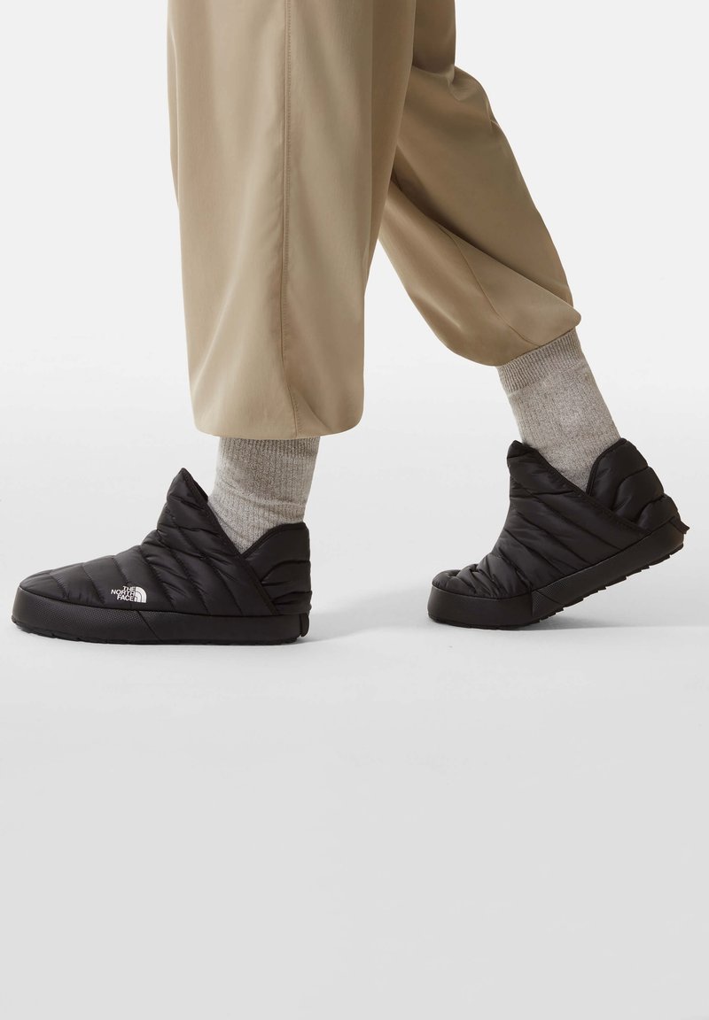 Personne portant un pantalon beige et des chaussettes grises avec des chaussures à enfiler matelassées noires de The North Face marchant sur un fond blanc.