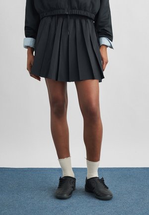 Persoon die een zwarte geplooide rok draagt, een zwart jasje met opgerolde mouwen, witte sokken en zwarte sneakers, staand op een blauw tapijt.