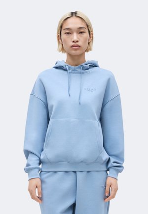 Νεαρό άτομο με κοντά ξανθά μαλλιά που φοράει γαλάζιο φούτερ με κουκούλα και αντίστοιχο παντελόνι, στέκεται μπροστά σε απλό φόντο.