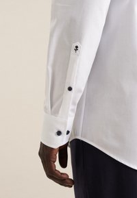 Mørkhudet hånd hviler ved siden af kroppen iført en hvid skjorte med sorte knapper og en lille sort rosebroderi på ærmet.