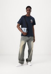 Camiseta de color azul marino con gráfico en el pecho, bolsa clara con correa negra, jeans gris claros y zapatillas blancas. Atuendo sencillo y casual.