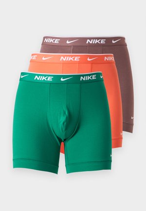 Homme torse nu, debout pieds nus, portant un boxer marron Nike, regardant légèrement de côté sur un fond clair uni.
