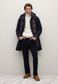 Cappotto blu navy con cappuccio, maglione color crema a trecce, sciarpa a quadri, pantaloni neri e scarpe color cammello. Tessuto liscio e silhouette strutturata.