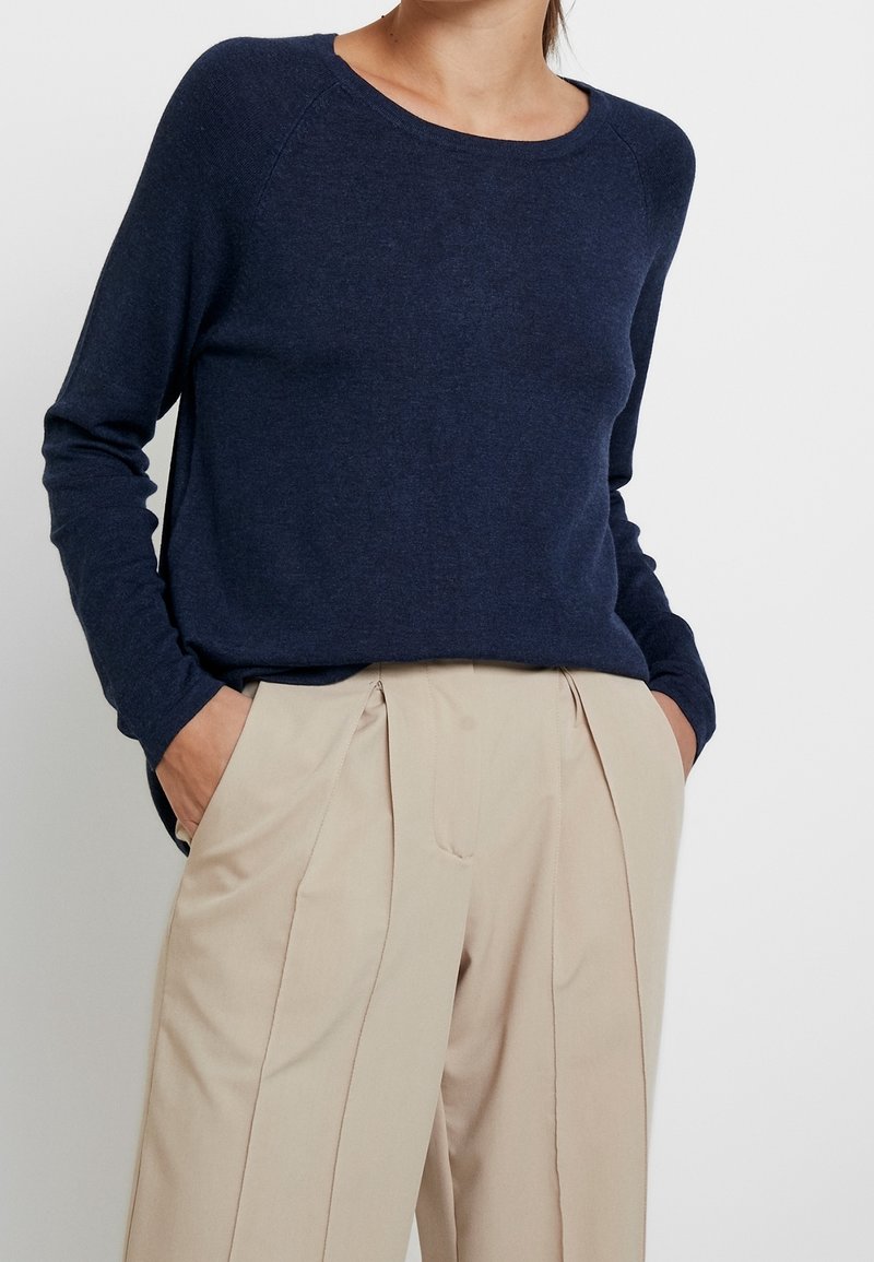 Maglione a maglia blu navy con maniche lunghe e scollo rotondo, abbinato a pantaloni beige a pieghe. Tessuto liscio e vestibilità comoda.