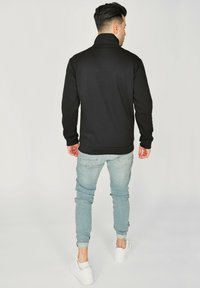 Czarna kurtka zapinana na zamek z wysokim kołnierzem, miękką teksturą i długimi rękawami, noszona na jasnoniebieskich dopasowanych dżinsach i białych trampkach.