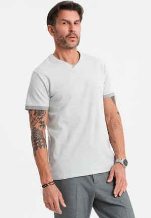 Camiseta blanca de cuello en V con mangas cortas, con ribete gris en el escote y las mangas. Tejido cómodo. El modelo lleva pantalones grises y accesorios.