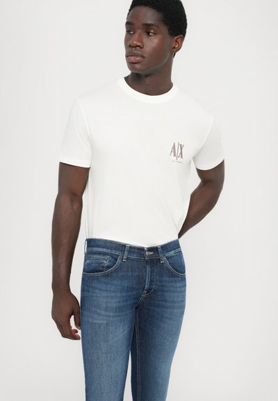 Vit t-shirt med rund halsringning och en liten brun logotyp på bröstet, ihop med mörkblå denimjeans med klassisk femficksdesign.